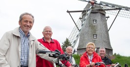 Das Bild zeigt eine Gruppe von Menschen auf Fahrrädern. Im Hintergrund steht eine Windmühle.